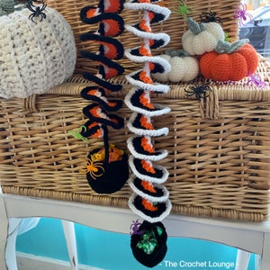 May include: Crocheted Halloween decorations featuring spiral garlands in black, orange, and white, along with crocheted pumpkins and black cauldron candy holders. The decorations are displayed on a wicker basket, creating a festive autumn scene.