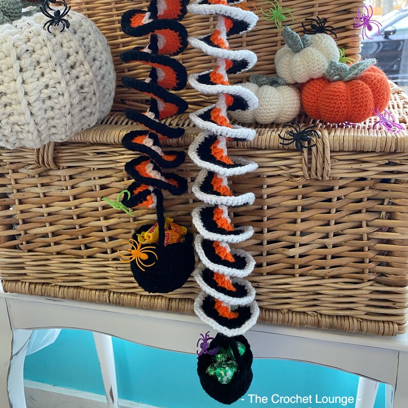 May include: Crocheted Halloween decorations featuring spiral garlands in black, orange, and white, along with crocheted pumpkins and black cauldron candy holders. The decorations are displayed on a wicker basket, creating a festive autumn scene.