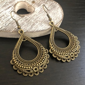 May include: A pair of gold-toned dangle earrings with intricate, detailed designs. The earrings are shaped like teardrops with a large, open circle in the center.