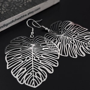 May include: Silver earrings shaped like monstera leaves. The leaves are cut out and have a delicate, intricate design.