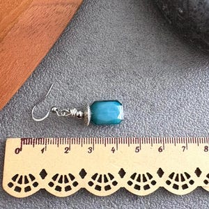 May include: A single earring featuring a teal, rectangular gemstone. The earring has a silver hook and decorative metalwork. The gemstone is approximately 2 cm long. The earring is displayed on a gray surface.