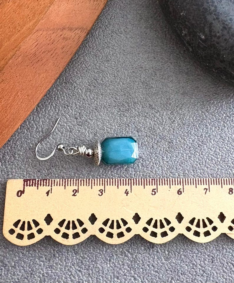 May include: A single earring featuring a teal, rectangular gemstone. The earring has a silver hook and decorative metalwork. The gemstone is approximately 2 cm long. The earring is displayed on a gray surface.