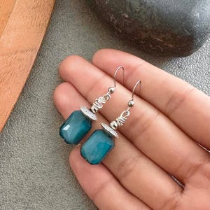May include: Teal gemstone earrings with silver detailing. The earrings have a rectangular, faceted design and are suspended from silver hooks. The silver elements include wire wrapping and decorative caps. The earrings are a striking accessory.
