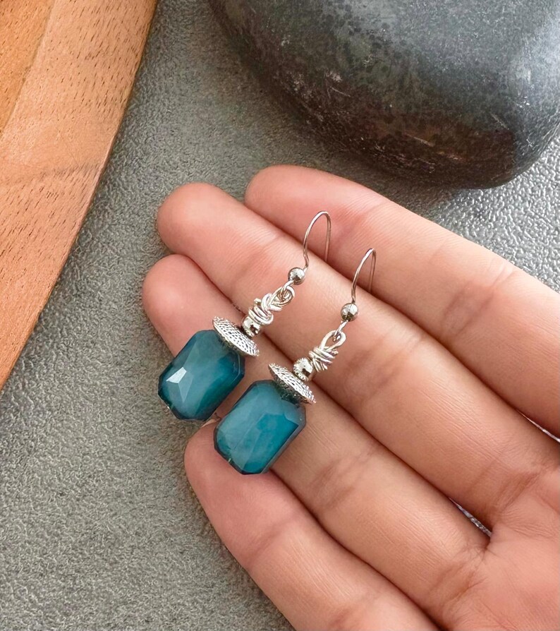 May include: Teal gemstone earrings with silver detailing. The earrings have a rectangular, faceted design and are suspended from silver hooks. The silver elements include wire wrapping and decorative caps. The earrings are a striking accessory.