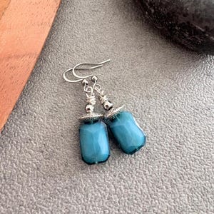 May include: A pair of teal-coloured, faceted glass earrings with silver-tone metal accents. The earrings have a rectangular shape with rounded edges and are suspended from silver-tone ear wires. The earrings are displayed on a textured grey surface.