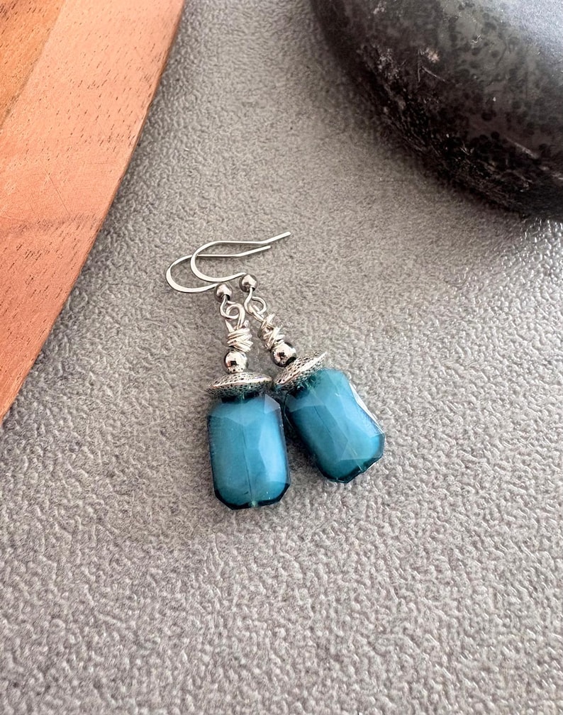 May include: A pair of teal-coloured, faceted glass earrings with silver-tone metal accents. The earrings have a rectangular shape with rounded edges and are suspended from silver-tone ear wires. The earrings are displayed on a textured grey surface.