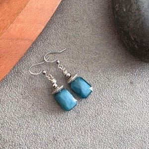 May include: Teal-coloured, faceted gemstone earrings with silver metal detailing. The rectangular gemstones are suspended from silver ear wires. The earrings are displayed on a textured grey surface. The gemstones have a polished, reflective surface.