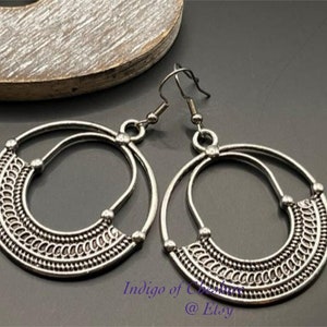 May include: A pair of silver hoop earrings with a detailed, intricate design. The earrings have a circular shape with a smaller circle inside. The earrings are made of metal and have a shiny finish.