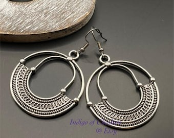 May include: A pair of silver hoop earrings with a detailed, intricate design. The earrings have a circular shape with a smaller circle inside. The earrings are made of metal and have a shiny finish.