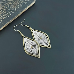 May include: Two teardrop earrings with a filigree design. The earrings have a gold-coloured frame and a silver-coloured, woven interior. A small gold bead is in the centre. The earrings are displayed on a dark surface.