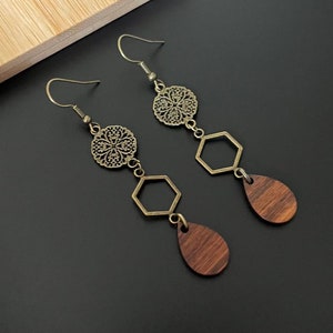 May include: Long drop earrings with a vintage design. Each earring has a filigree circle, a hexagonal shape, and a teardrop-shaped wooden pendant. The metal parts have an antique brass finish, set against a black background.