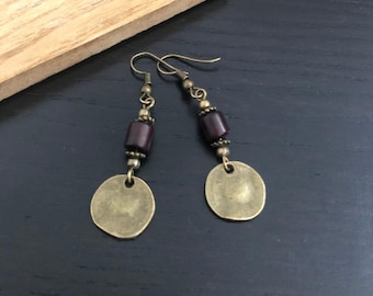 May include: A pair of dangle earrings with a vintage aesthetic. Each earring features a bronze-colored disc, a dark red cylindrical bead, and smaller bronze beads. The earrings hang from bronze-colored ear wires.