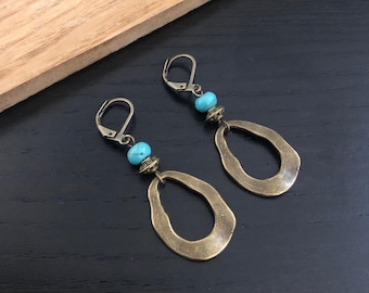 Puede incluir: Un par de pendientes colgantes con acabado bronce. Cada pendiente presenta una cuenta turquesa y un colgante abierto en forma de lágrima de bronce. Cierre de palanca. Fondo de superficie de madera oscura.