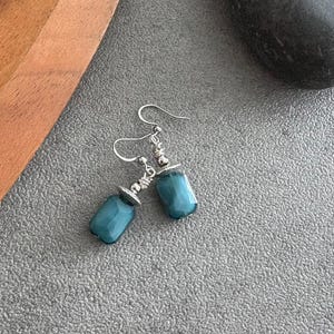 May include: A pair of teal earrings with silver hooks and decorative caps. The earrings have a rectangular, faceted bead design. The jewellery is displayed on a grey surface.