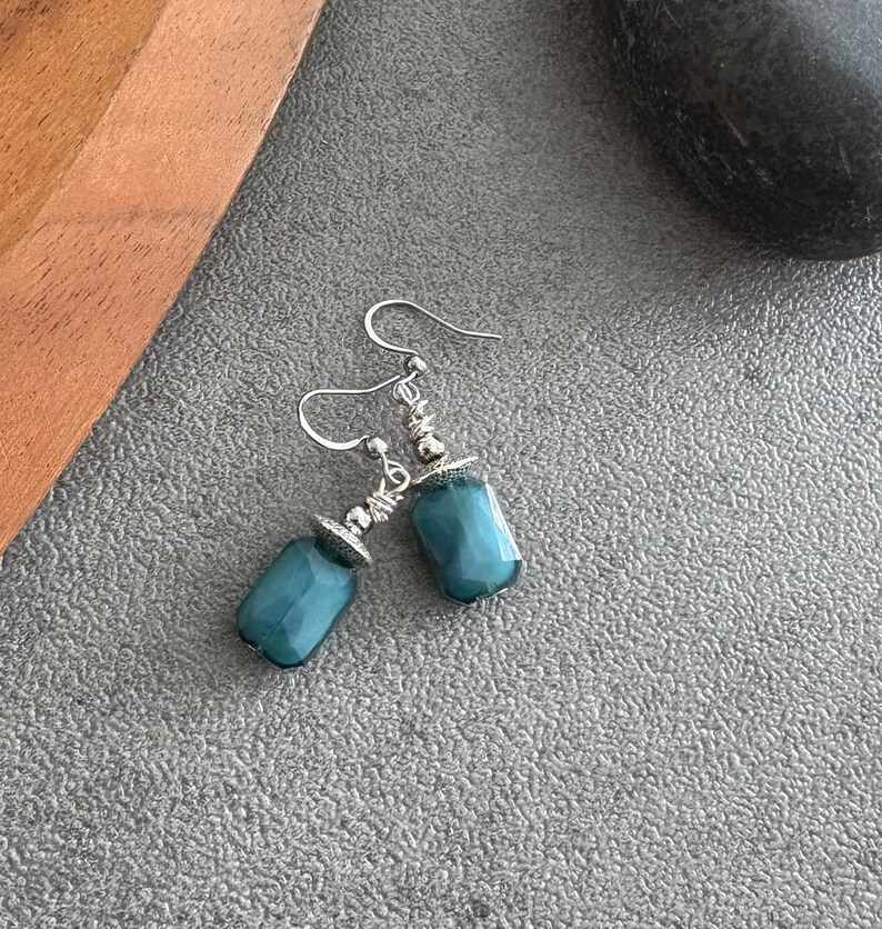 May include: A pair of teal earrings with silver hooks and decorative caps. The earrings have a rectangular, faceted bead design. The jewellery is displayed on a grey surface.