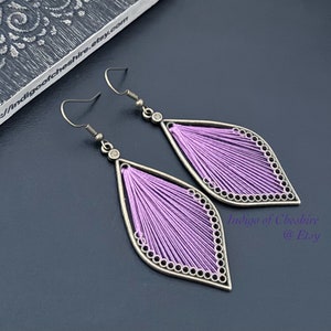 Peut inclure: Une paire de boucles d'oreilles pendantes de forme goutte. Les boucles d'oreilles sont en métal argenté et présentent un motif délicat et tissé de fil lavande. Les boucles d'oreilles sont rehaussées d'un petit strass transparent.