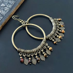 May include: A pair of antique gold-coloured hoop earrings. The earrings have detailed leaf-shaped charms and small, round beads in shades of brown and red. The earrings are displayed on a dark surface.