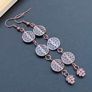 May include: A pair of long drop earrings featuring a copper-toned finish with a hammered, textured surface. Each earring consists of three circular discs connected by small metal rings, ending with a small leaf-shaped charm at the bottom.