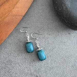 May include: A pair of turquoise-coloured, rectangular bead dangle earrings. The earrings have silver-coloured hooks and decorative accents. The beads are a vibrant shade of blue-green. The earrings are displayed on a grey surface.