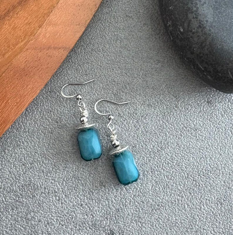 May include: A pair of turquoise-coloured, rectangular bead dangle earrings. The earrings have silver-coloured hooks and decorative accents. The beads are a vibrant shade of blue-green. The earrings are displayed on a grey surface.