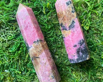 Puede incluir: Dos puntas de piedras preciosas pulidas, también conocidas como rodonita, en tonos rosados y marrones, sobre un lecho de musgo verde.