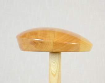 May include: A wooden hat form with a smooth, rounded top and a cylindrical base. The form is made of light brown wood and has a natural finish.