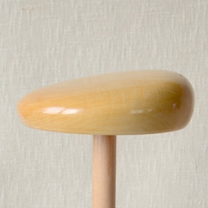 Può includere: Un forma per cappello in legno con una parte superiore arrotondata liscia e una base cilindrica. La forma è di colore marrone chiaro e sembra essere in legno grezzo.