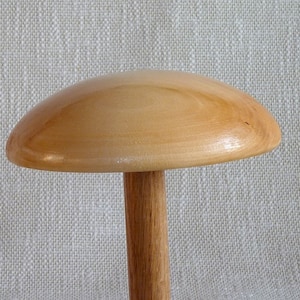 Op de afbeelding: Een houten hoedstandaard in de vorm van een paddenstoel, met een lichtbruine afwerking. De standaard heeft een gladde, ronde bovenkant en een slanke, cilindrische basis.