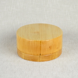May include: A round wooden box with a natural wood finish. The box has a lid and is made of light-colored wood.
