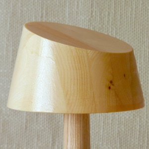 May include: A wooden lamp with a light brown shade and a cylindrical base. The shade is angled and has a smooth, polished finish.