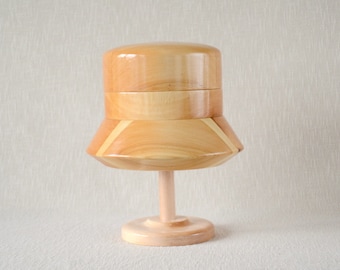 May include: A wooden hat form with a light brown finish. The form is made of multiple pieces of wood that are joined together. The form is on a wooden stand.