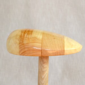 Op de afbeelding: Een houten hoedblok met een lichtbruine afwerking. Het blok is gemaakt van meerdere stukken hout die aan elkaar zijn gelijmd. Het blok is gevormd als een hoed en heeft een ronde bovenkant en een smalle basis.