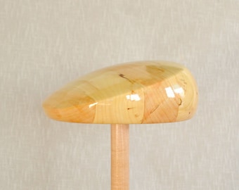 May include: A wooden hat block with a light brown finish. The block is shaped like a traditional cap and has a smooth, polished surface.