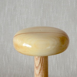 Op de afbeelding: Een houten garenwinder in de vorm van een paddenstoel, met een lichtbruine bovenkant en een donkerbruine basis.