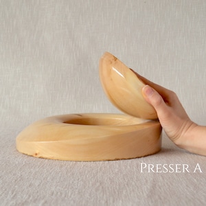 May include: A wooden hat presser with a lid that is being lifted open. The presser is made of light wood and has a smooth, rounded shape. The text "PRESSER A" is visible in the lower right corner of the image.