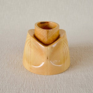 May include: A handcrafted wooden candle holder with a natural finish. The candle holder has a unique design, featuring a rounded base and a cylindrical top to hold a candle. The wood grain is visible, adding to its rustic charm.