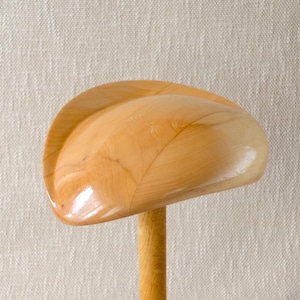Pode incluir: Um bloco de chapéu de madeira clara e lisa com um topo arredondado em forma de cúpula e uma base cilíndrica. O veio da madeira é visível em toda a superfície polida, mostrando variações naturais nos tons bege e castanho claro do material.