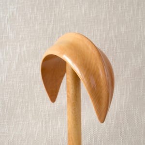 Op de afbeelding: Een houten hoedenblok, met een gebogen, hoedvormige vorm op een cilindrische houten paal. Het hoedenblok is gemaakt van lichtgekleurd hout, met een gladde, gepolijste afwerking. Het ontwerp is eenvoudig en elegant.