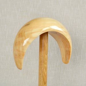 May include: A wooden hat stand with a curved, semi-circular top and a vertical support. The stand is made of light-colored wood with a glossy finish, showcasing the natural grain. The design is simple and functional, suitable for displaying hats.