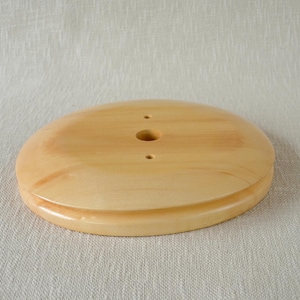 May include: A round, light-colored wooden base with a smooth, polished finish. The circular object features a larger central hole flanked by two smaller, symmetrical holes positioned vertically along the center line of the wood surface.