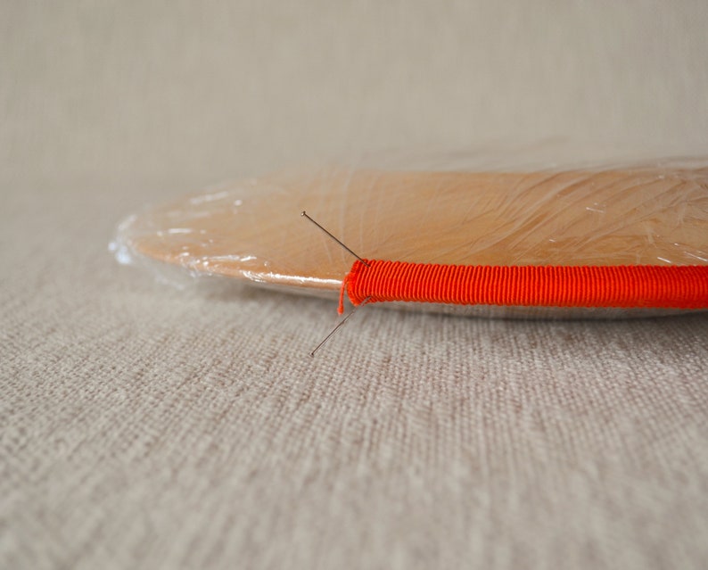 May include: A round wooden plate with a clear plastic wrap over it. A bright orange elastic band is wrapped around the edge of the plate and secured with two pins.