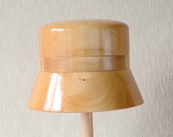 May include: A wooden hat form with a light brown finish. The form has a wide brim and a rounded crown.