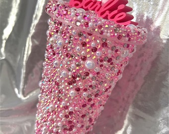 Op de afbeelding: Roze herbruikbare beker bedekt met roze en witte strass steentjes met een roze "Barbie" naamplaatje bovenop en een roze rietje.