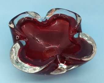 May include: A decorative, ruby red glass bowl with a unique, wavy shape. The bowl has a clear, silver-toned rim and a glossy finish. The bowl is set against a light blue background.