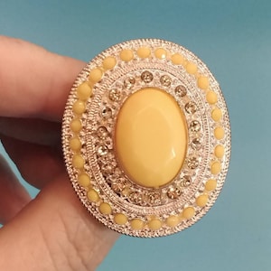 May include: A large, oval-shaped ring with a yellow stone set in a silver-toned metal frame. The frame is decorated with a circle of small yellow stones and a circle of clear rhinestones.