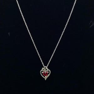Peut inclure: Collier argenté avec un pendentif en forme de cœur. Le pendentif présente une pierre rouge au centre, entourée d'un cadre argenté décoratif avec de petits accents clairs. Le collier est sur fond noir.