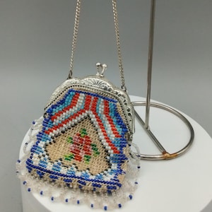 May include: A small, vintage beaded purse with a silver-tone frame and chain. The purse features a colorful geometric pattern in red, white, and blue, with a floral design in the center. The bottom edge has a fringe of clear beads.