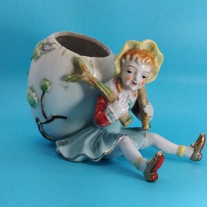May include: A porcelain figurine of a girl carrying a large, round planter. The planter is decorated with a floral design and the girl is wearing a blue dress, red jacket, and yellow hat.