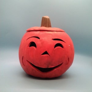 May include: A red ceramic pumpkin with a carved smiling face. The pumpkin has a brown stem and is a traditional Halloween decoration.
