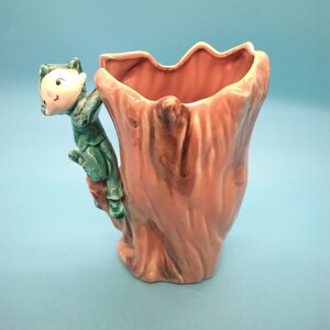 May include: A ceramic vase shaped like a tree trunk with a green and brown elf climbing up the side. The vase is a light brown colour with a pink glaze.